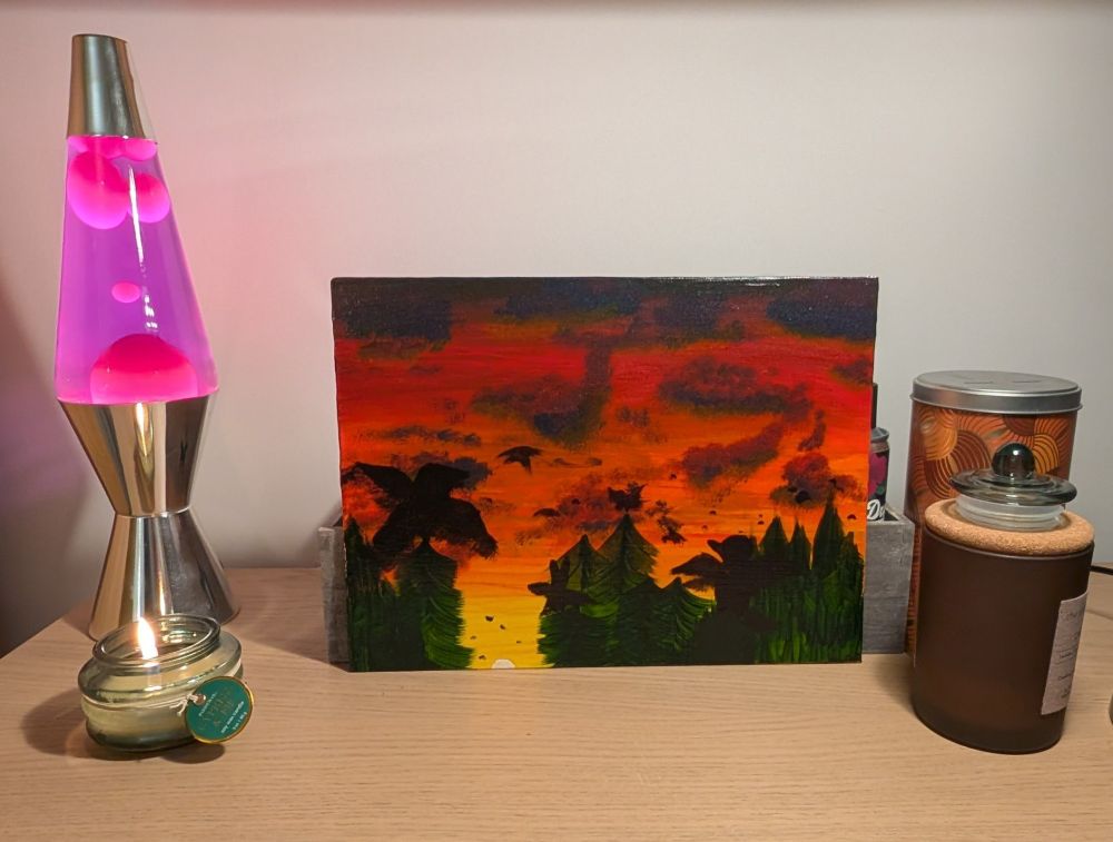 A painting of crows flying out of the woods at sunrise, around the painting are a lava lamp, a small green candle, a brown candle with a cork lid and the orange and silver tub that I keep my cotton buds in (cotton buds not pictured)