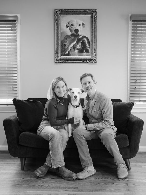 Adult couple sitting on the sofa with their beloved dog. Above them on the wall is a spoof oil painting of the dog as a medieval hero, named “Joan of Bark”.