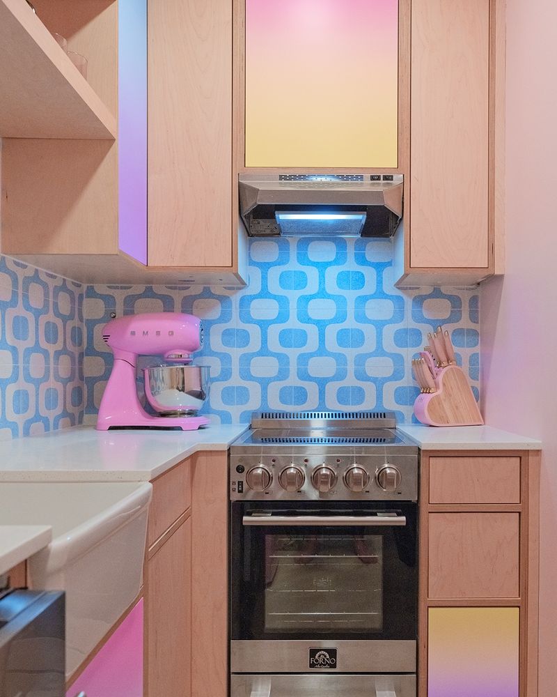A close-up view of a small, retro-inspired kitchen showcasing pastel gradient cabinet doors in pink, yellow, and blue. A pink stand mixer sits on the counter beside a modern stainless steel oven and hood. The backsplash features a bold blue geometric pattern, and light wood cabinetry ties the design together. A wooden knife block shaped like a heart adds a playful touch on the right-hand counter.