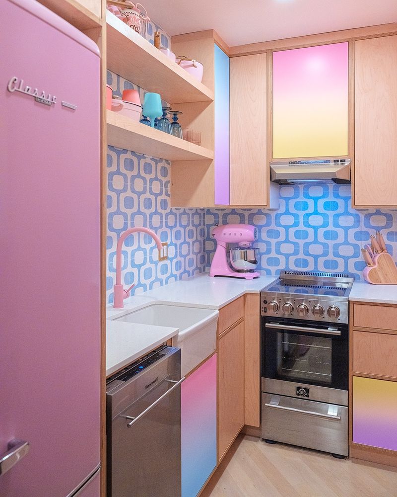 A vibrant, retro-inspired kitchen featuring pastel gradient cabinet doors in pink, yellow, and blue. A pink refrigerator and matching pastel pink faucet complement the design, along with a pink stand mixer on the counter. The backsplash has a blue geometric pattern, and light wood cabinetry adds warmth. The compact space feels colorful, playful, and stylish.