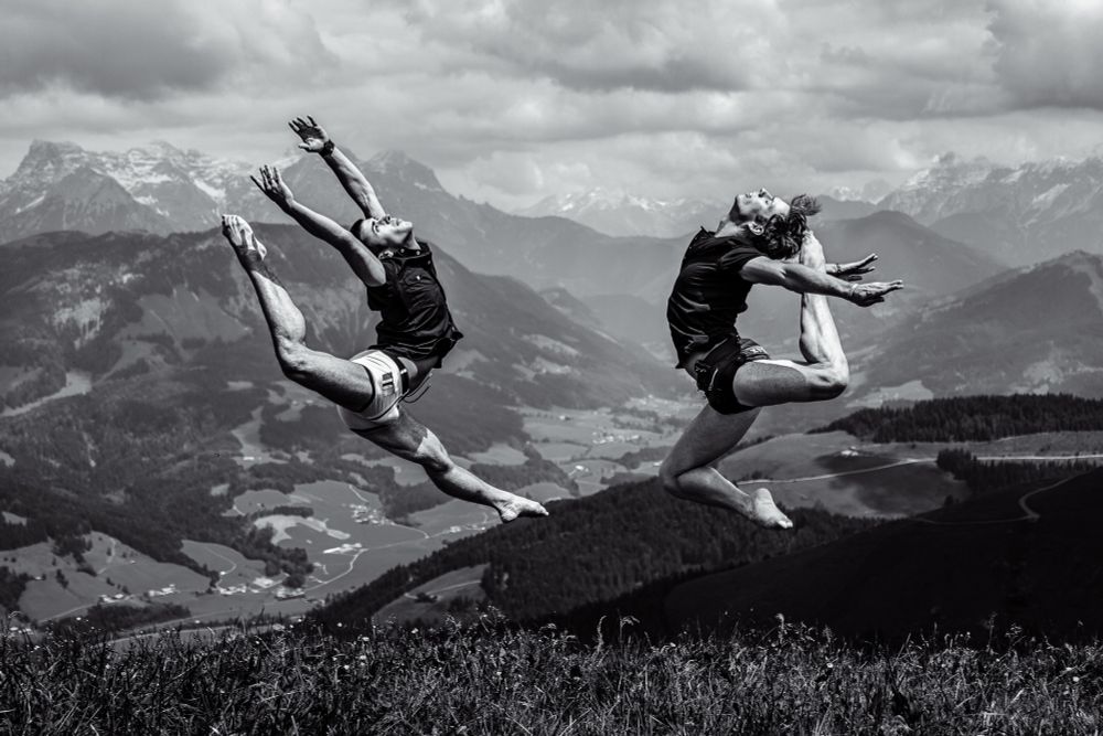 two male dancers leap into the air with the Austrian mountains in the background.