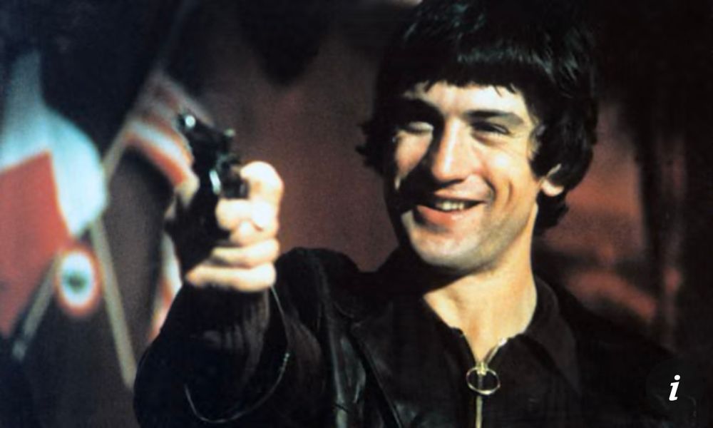 Screen still of a young Robert de Niro in one of Martin Scorcese’s early masterpieces, Mean Streets. De Niro is pointing a gun at someone. He has a smile on his face. It is all rather sinister 