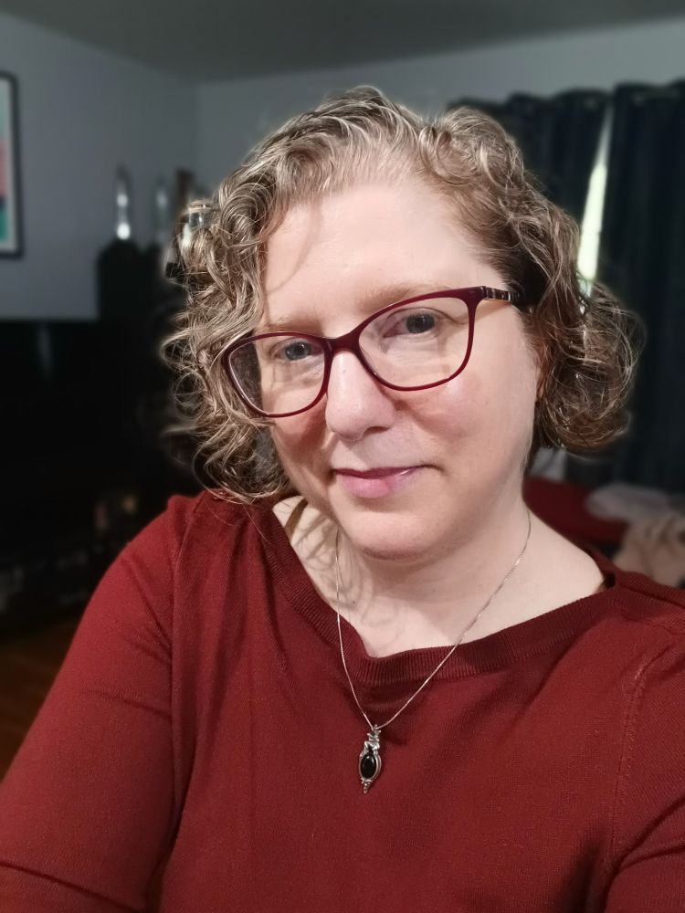 A middle aged woman with chin length curly hair wearing a Burgundy boatneck sweater and glasses with Burgundy frames.