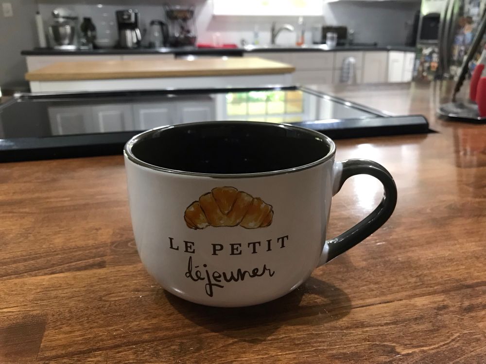 A round cappuccino mug that is dark brown on the inside, white on the outside, with a picture of a croissant and the words “le petit dejeuner” on the side.