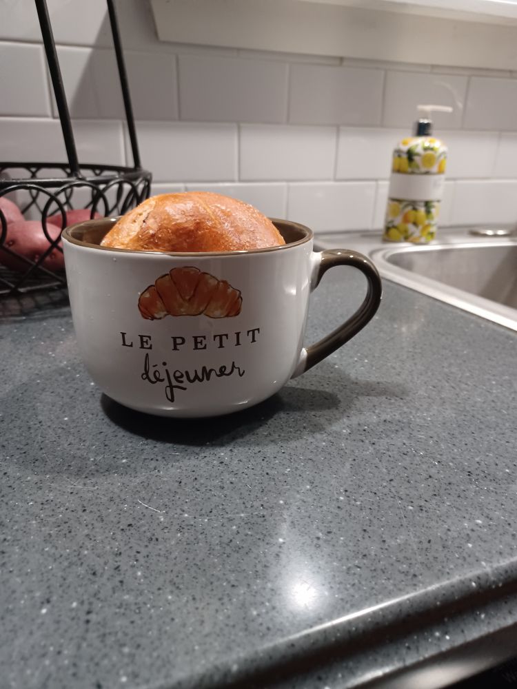 A white bowl-style mug with a brown handle and interior that has a picture of a croissant and says "le petit déjeuner." There is a raisin bagel propped inside it for scale.