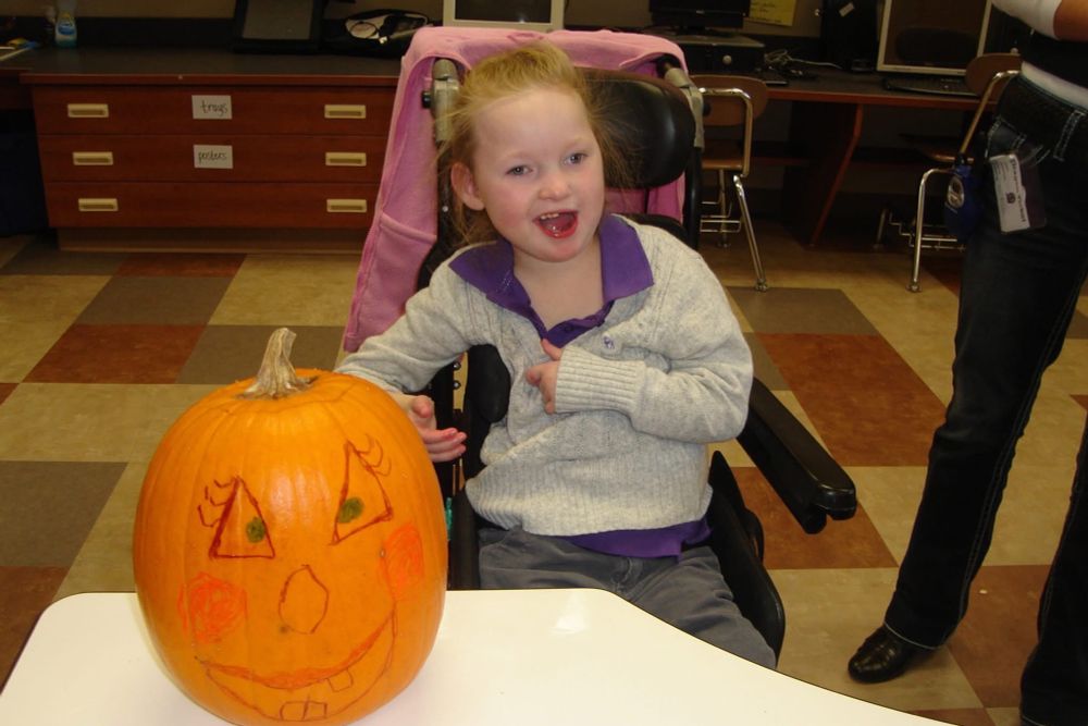 A blonde girl in a wheelchair wearing a gray sweater with a purple shirt underneath it smiles. There is a decorated pumpkin in the foreground. 