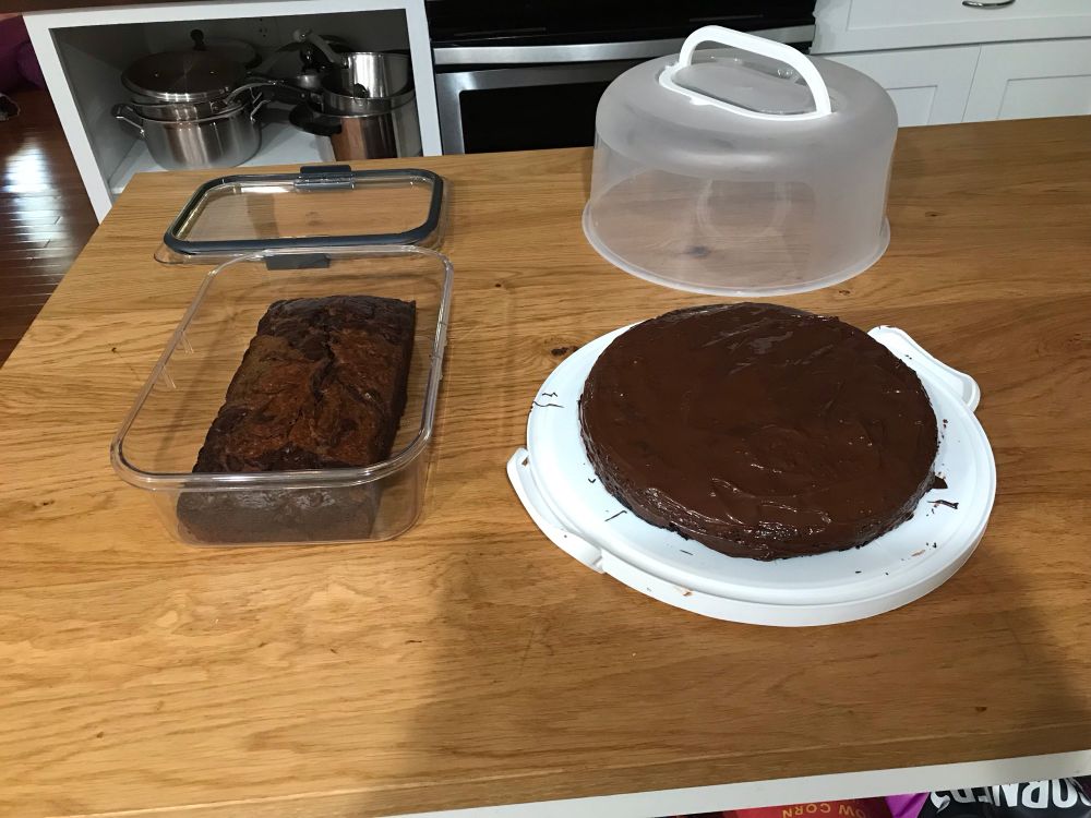 On the left, a banana bread in a rectangular container. On the right, a ganache covered round cake on a cake carrier base. 