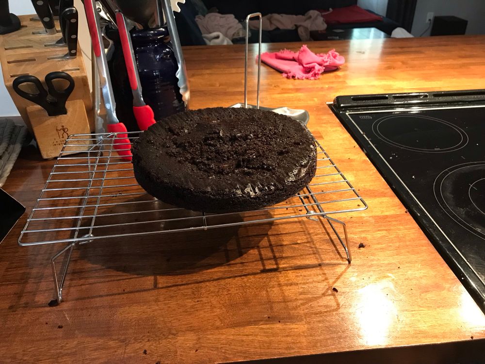 A flat round chocolate cake sits on a cooling rack.