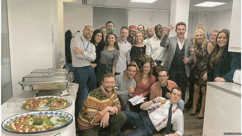Photo shows a group of people posing in a room, some with glasses raised, and buffet on table to side. 