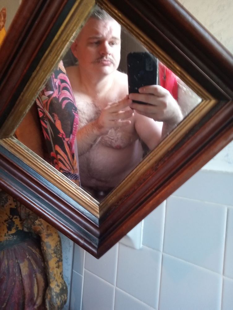 Antique frame hanging off a shower curtain rod with a bearish guy in the reflection, just a glint of metal cock ring