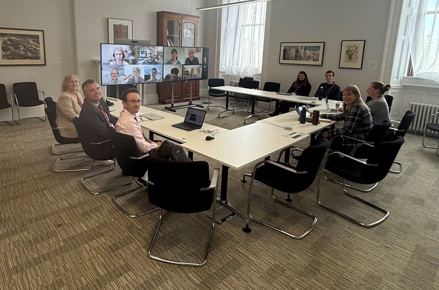 A group of 7 BAT stakeholders sat around a meeting table, with a wide video conferencing screen in the background with more online attendees