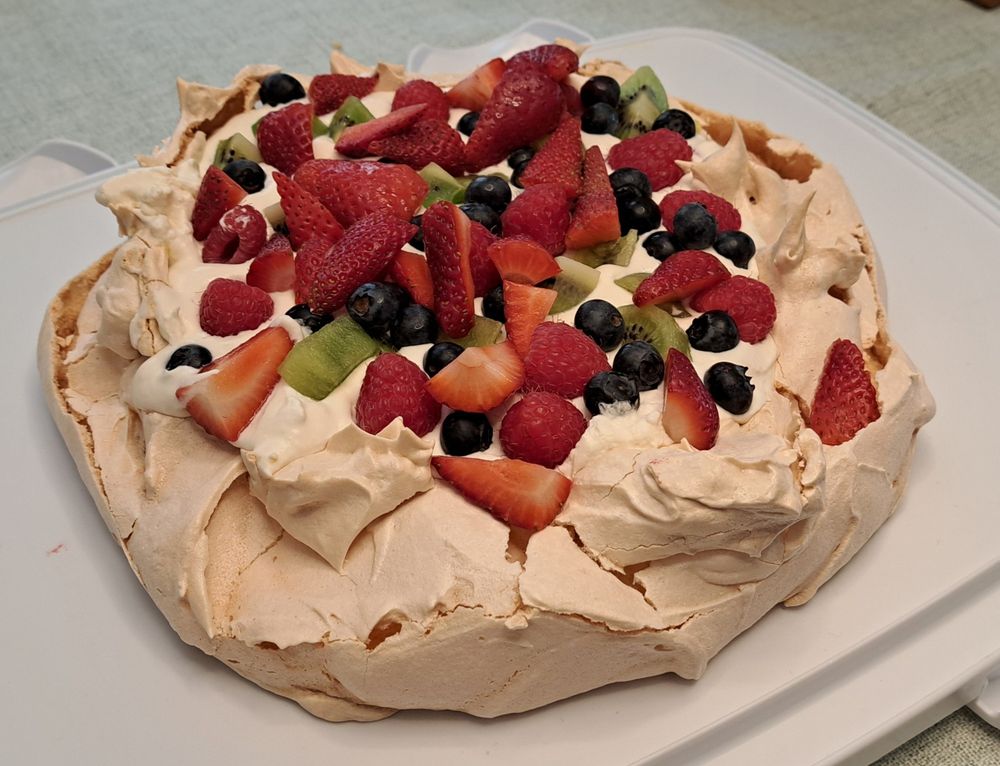 Pavlova covered in kiwi, strawberries, raspberries, and blueberries. There is a lemon curd beneath the cream.