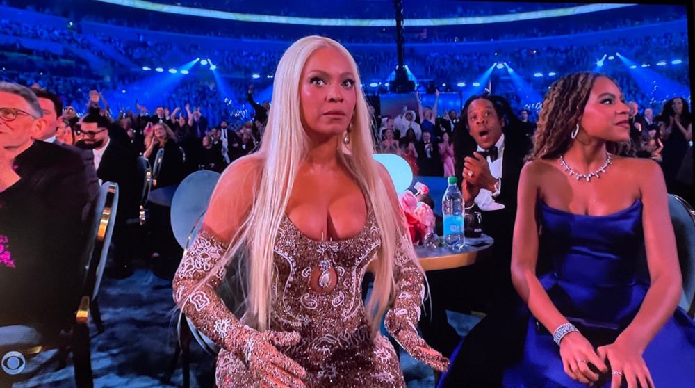 Beyoncé at the Grammys when Cowboy Carter won. She has her arms opened at her sides, her eyes are wide in shock, and her mouth is closed. She wears a sparkly dress and a long, platinum blonde wig. Blue Ivy is seated to her left in a blue dress. Jay Z is behind them, applauding, with a visibly shocked look on his face, jaw dropped.