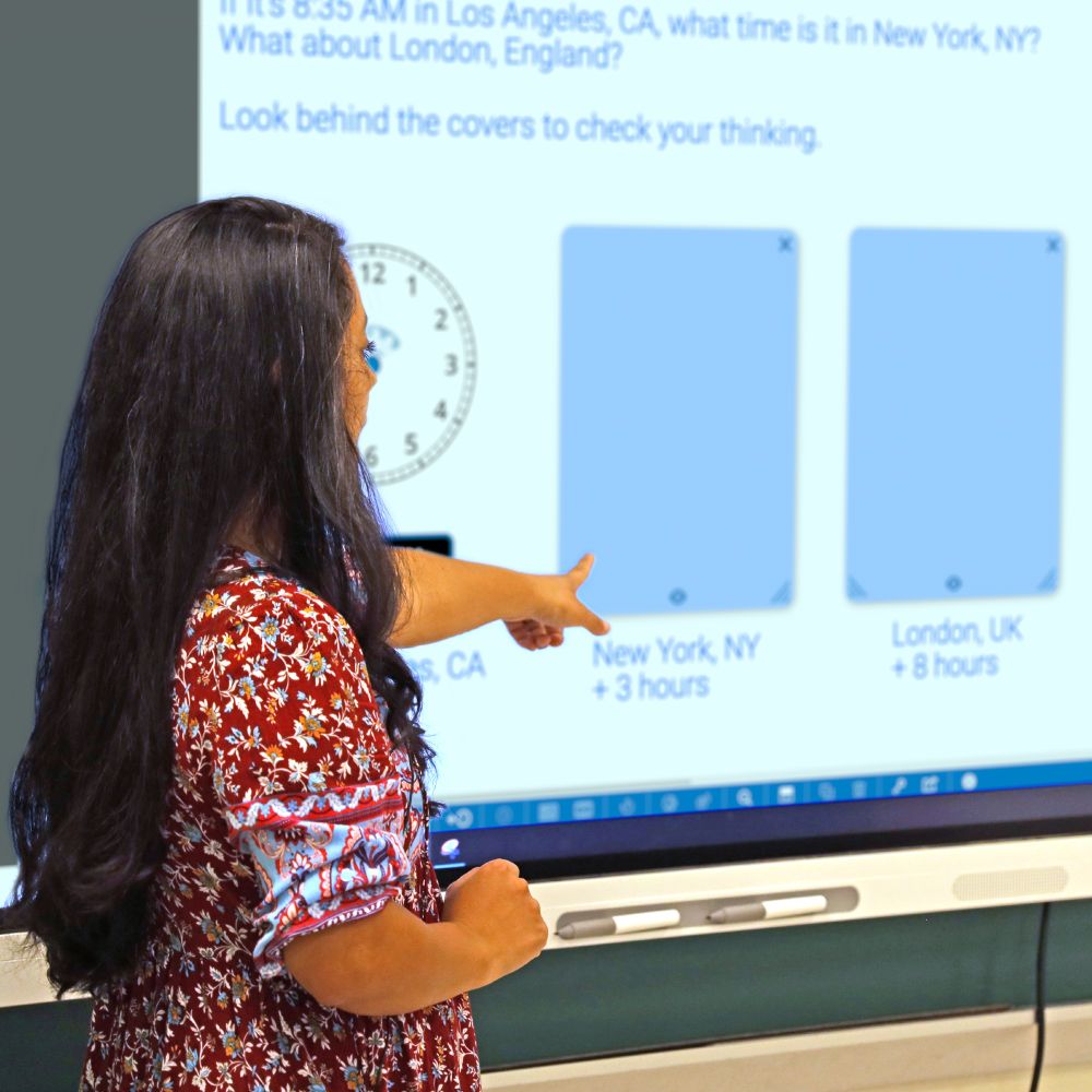 A teacher presenting a lesson, pointing to a digital display of the math clock app.