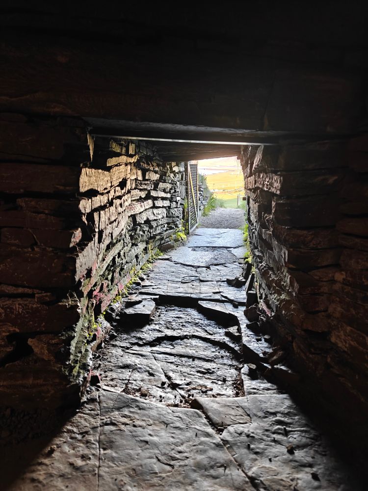 Narrow crawling tunnel with thinly layered stones. View is out the tunnel from inside an ancient chamber toward the sunny outdoors.