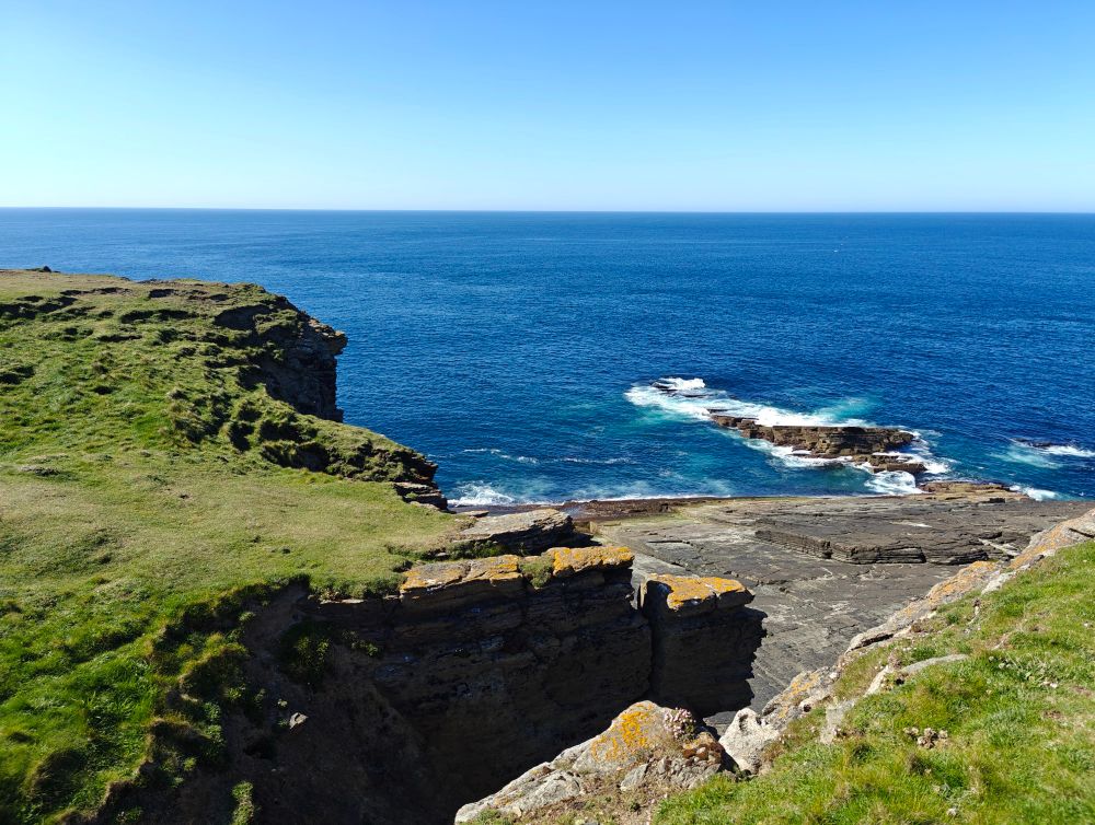 Oceanside view from the top of a cliff, showing water breaking on the rocks below, bare rocks on the cliff side and green grass on the crest.