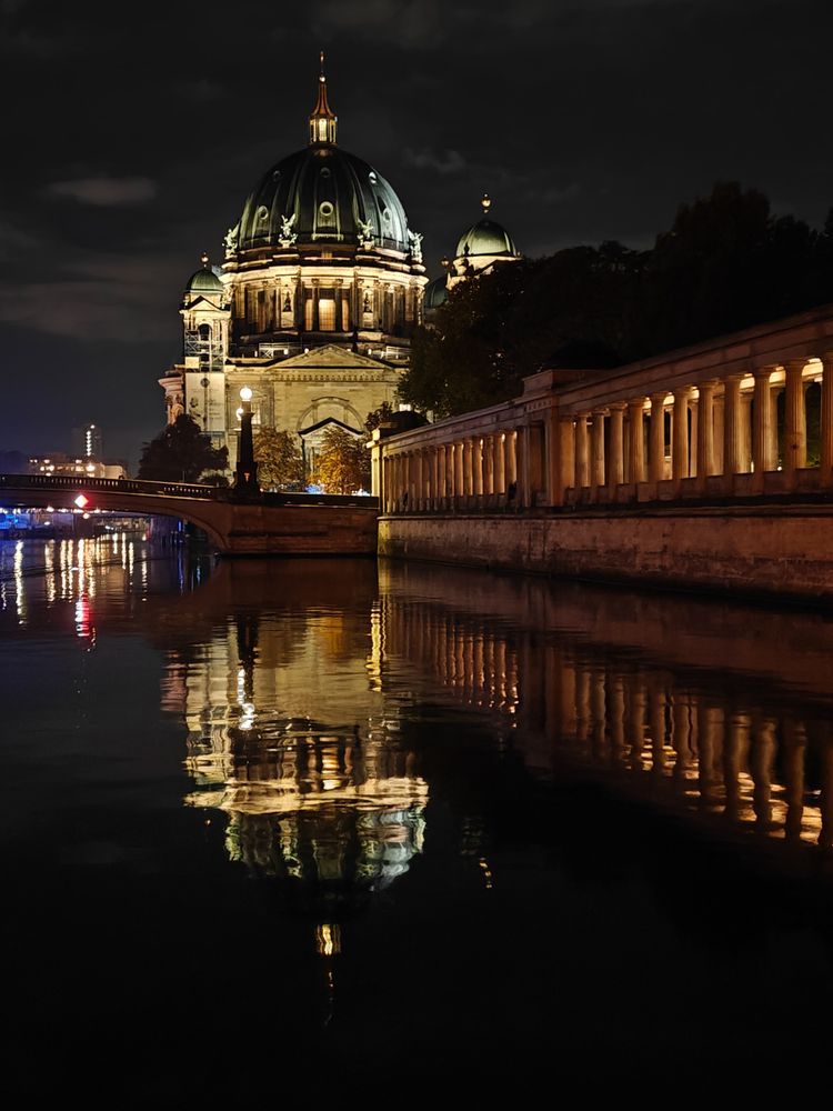 Nachtaufnahme der Museumsinsel in Berlin mit dem Dom und dem Säulengang der Alten Nationalgalerie daneben. Die Gebäude sind beleuchtet und der Dom spiegelt sich im Wasser der Spree.