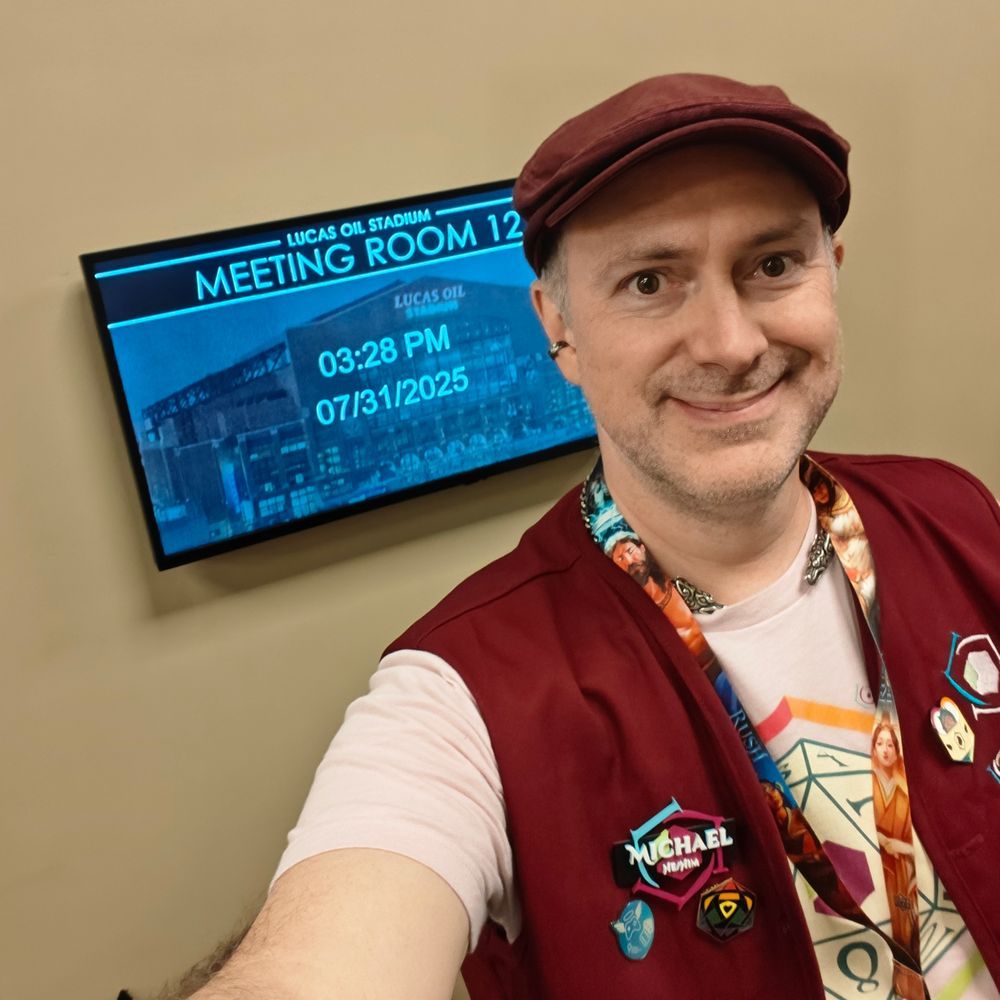 Michael wearing burgundy color Lore Link gear, flat cap, and name tag, standing in front of Meeting Room 12.