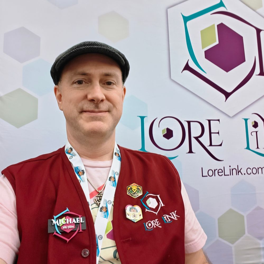 Michael wearing a pink Lore Link T-shirt, flat cap, maroon red vest, a large name tag with he/him pronouns, and a couple large pins on the vest.