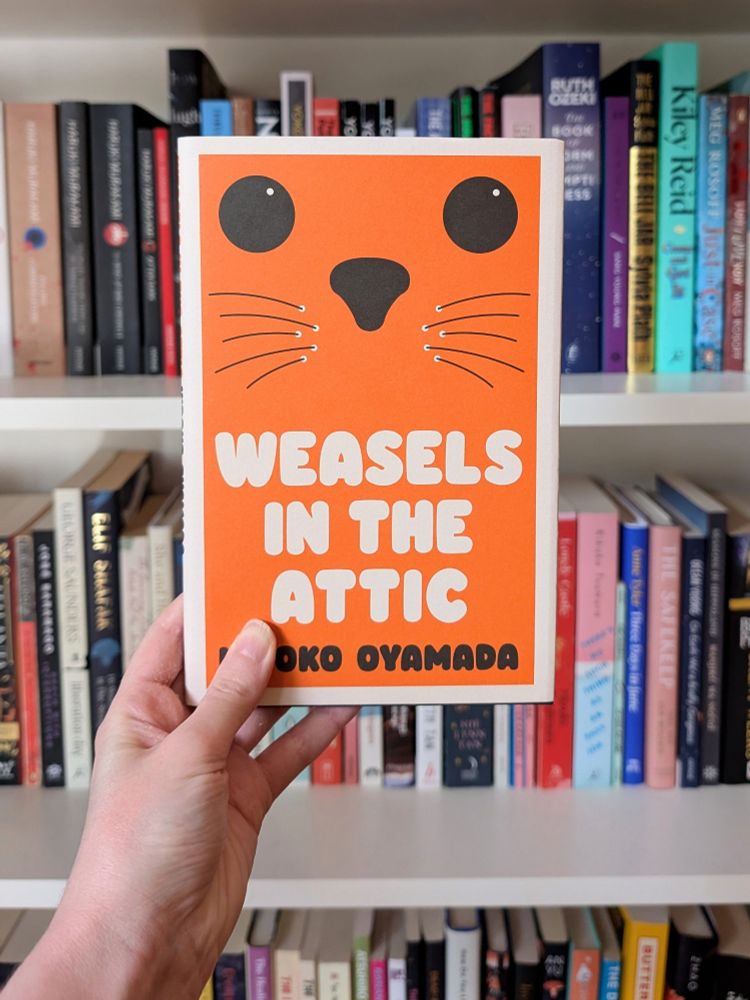Hand holding book Weasels in the Attic by Hiroko Oyamada in front of bookcase 