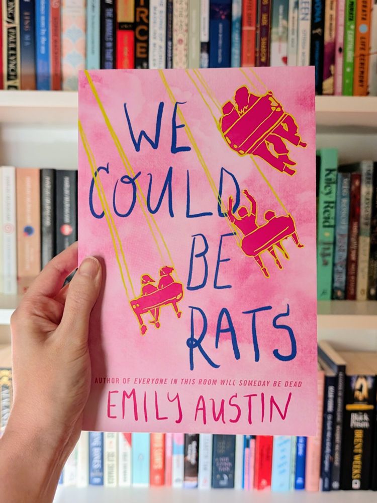 Hand holding up the book 'We Could Be Rats' by Emily Austin in front of a bookcase 