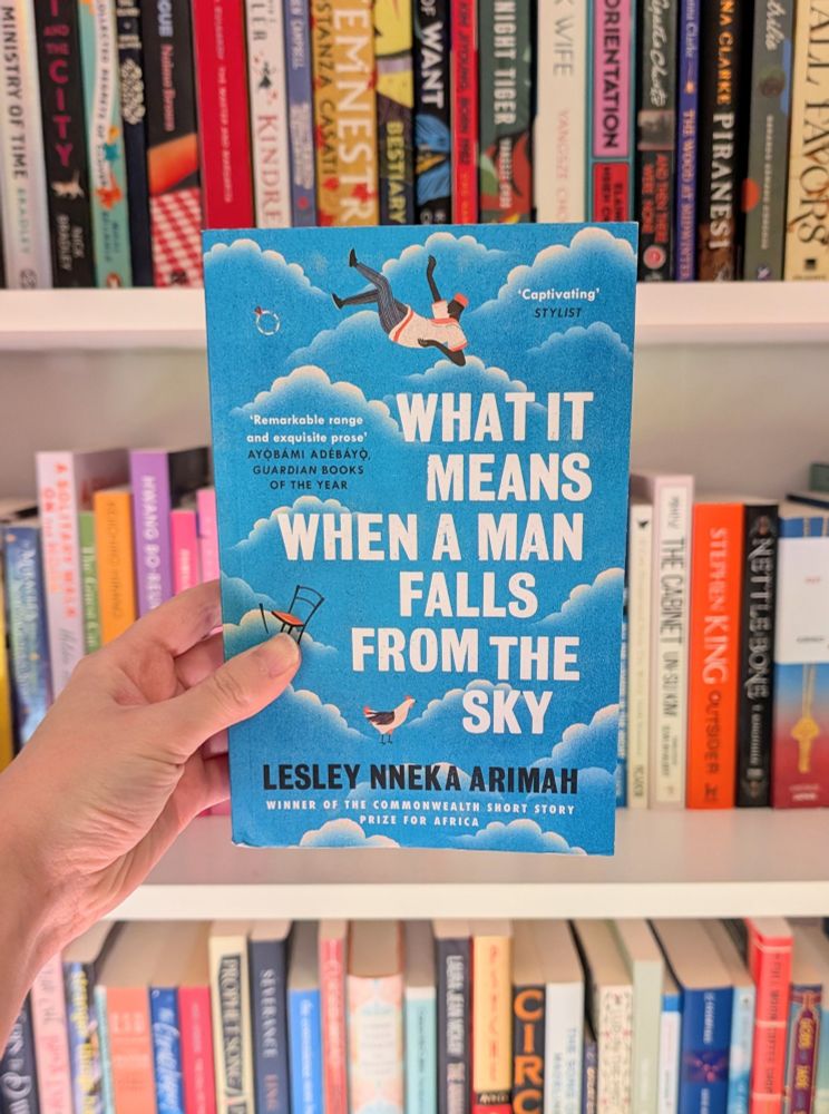 Hand holding up a book in front of a bookcase. The book is What It Means When A Man Falls From The Sky by Lesley Nneka Arimah