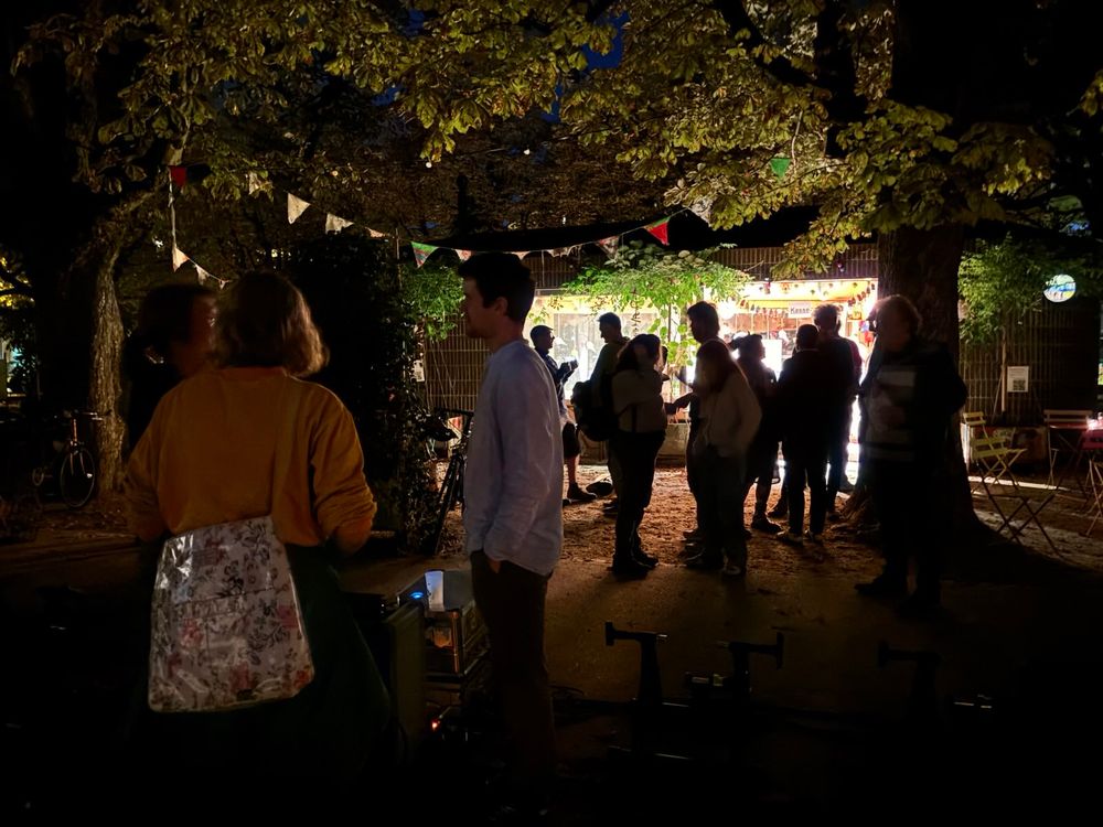 Menschen stehen im Dunklen und sprechen miteinander unter Bäumen vor einer hell erleuchteten Bar.