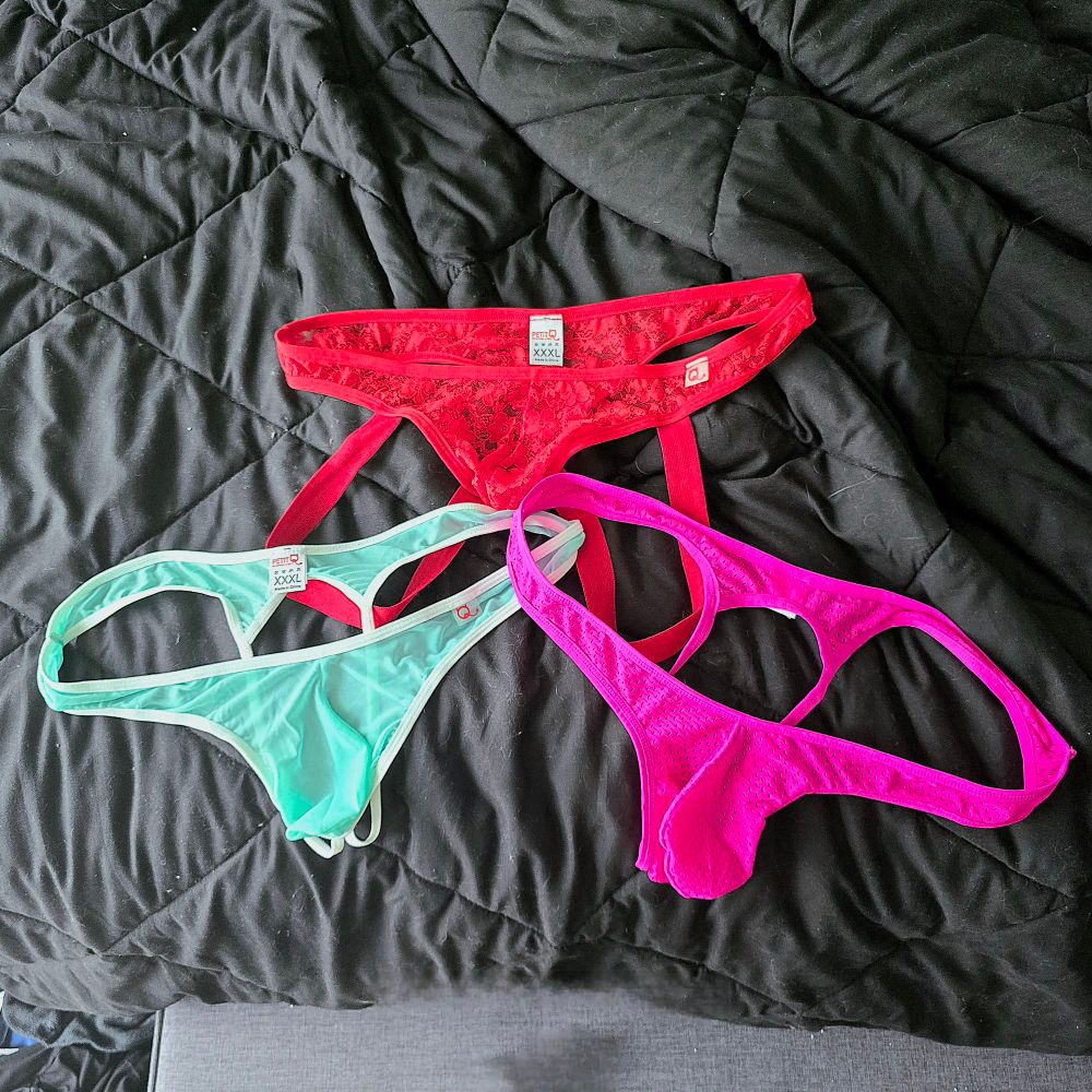 A red jock thong, a seafoam green thong, and a pink thong on top of a black bed comforter