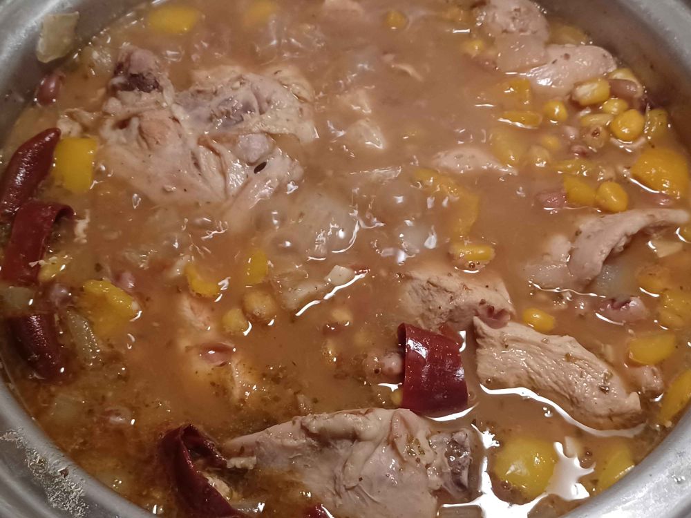 closeup of a pot of soup with visible chunks of chicken, dent corn, anasazi beans, onions, gold bell pepper, guajillo chile, and seasonings simmering away.