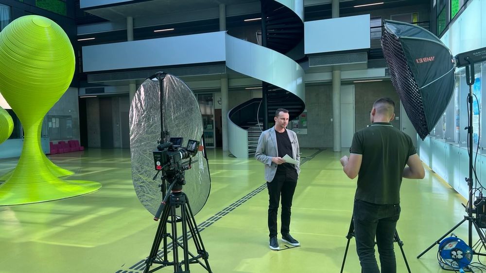 Florian Tschorsch steht in einem modernen Gebäude mit grünem Boden vor der Kamera, während ein Kamerateam mit Lichtequipment und Reflektor eine Videoaufnahme vorbereitet. Im Hintergrund ist eine spiralartige Treppe zu sehen sowie eine futuristische, grüne Skulptur.