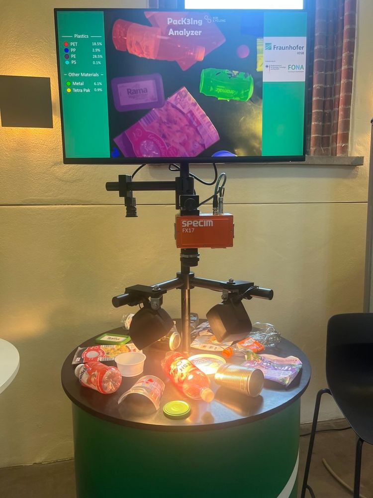 Ein Packing Alayzer im Ausstellungsraum. Er sieht aus wie ein runder Tisch mit Kunststoffverpackungen darauf, über dem eine verkabelte Vorrichtung installiert ist.