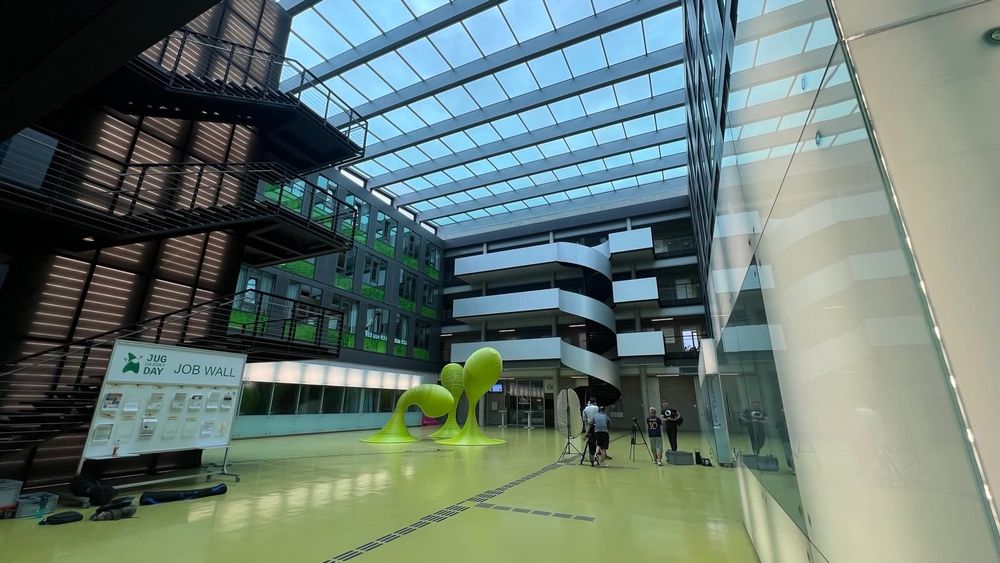 Weitwinkelaufnahme eines lichtdurchfluteten Atriums mit Glasdach und mehreren Etagen. In der Mitte des Raums befinden sich auffällige, grüne Skulpturen. Ein Kamerateam filmt Florian Tschorsch, während ein "Job Wall"-Board auf der linken Seite sichtbar ist. Moderne Architektur und klare Linien prägen die Umgebung.