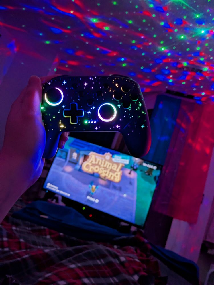 a photo of someone holding a rainbow led sparkling switch controller in front of their tv screen displaying animal crossing new horizons at the foot of a bed. the back wall has a bisexual pride flag hung up and a galaxy projector is on with the same colors as the flag, making the entire room a purpley color.
