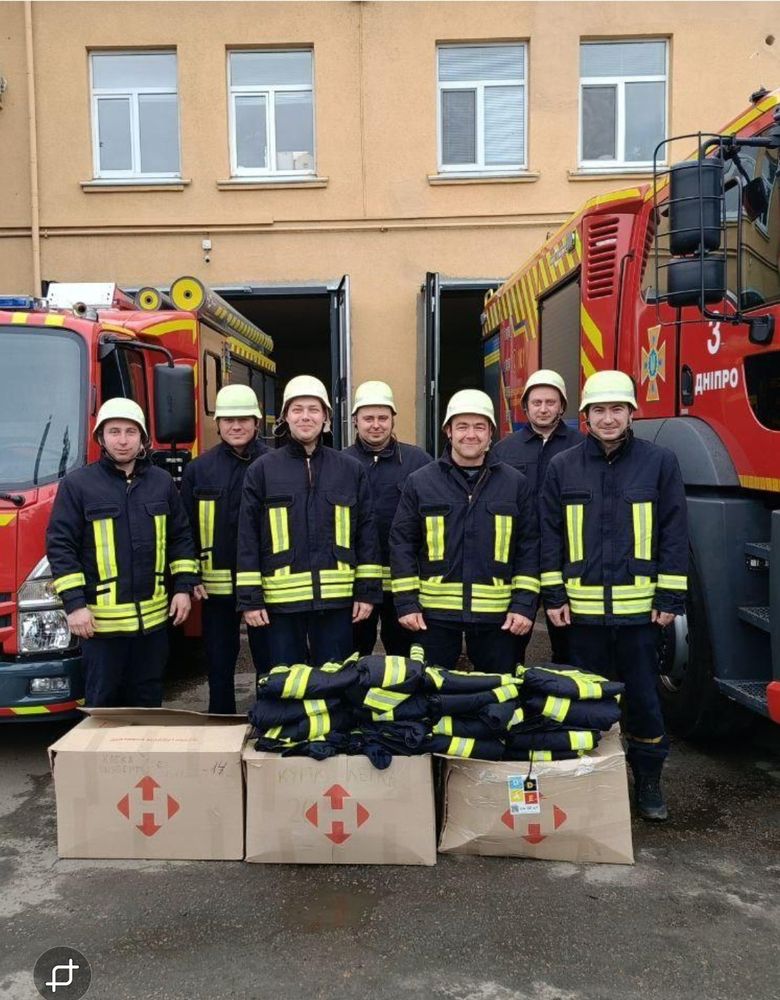 The photo shows 7 Ukrainian firefighters with two fire engines in the background and boxes in the foreground with a UA-DE sticker.