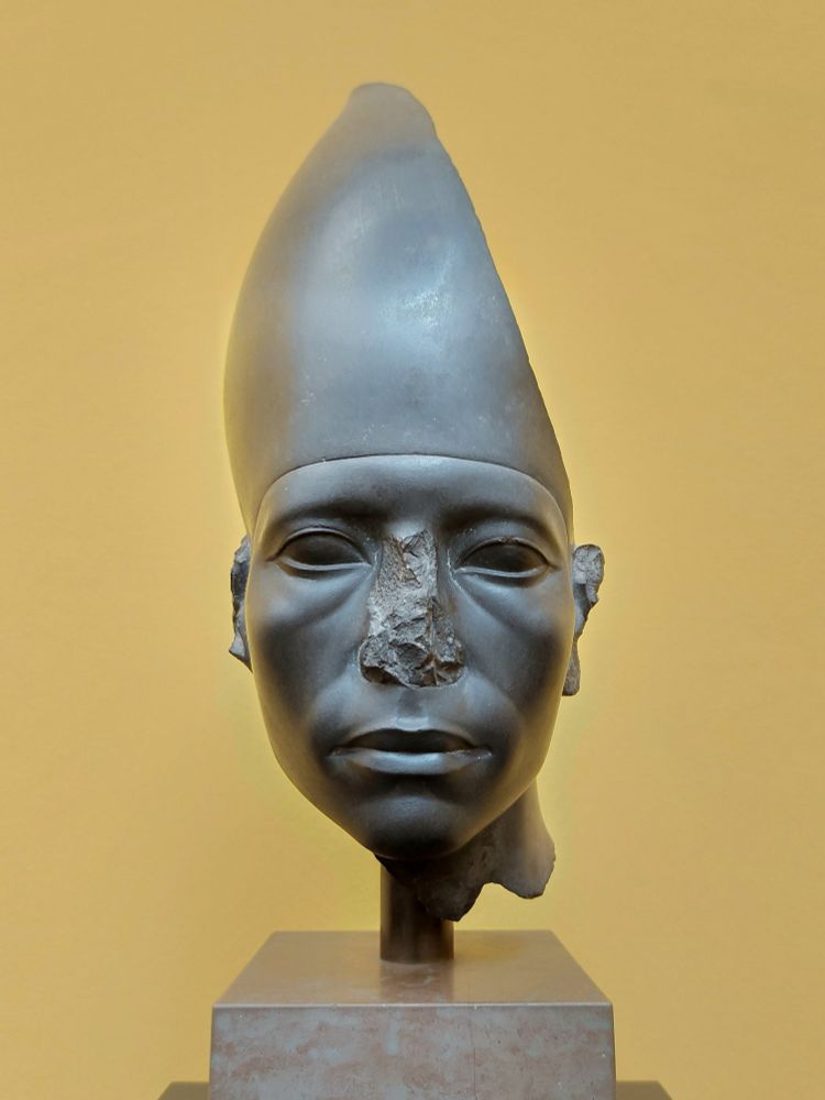 A portrait of a pharaoh, made of dark grey stone, displayed on a pedestal against a yellow background. The sculpture features a tall, conical White Crown of Upper Egypt. The face is well-defined but has visible damage, particularly on the nose and parts of the ears. Despite the missing details, the lips, eyes, and overall structure retain a striking presence