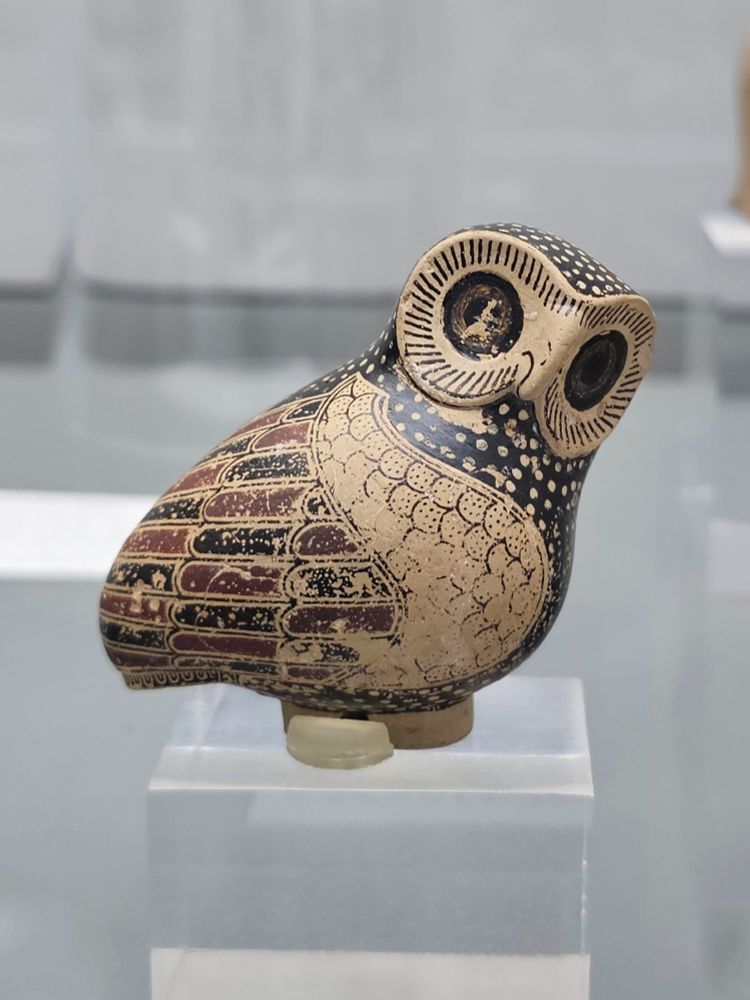 
A pottery owl figurine displayed on a transparent pedestal. The figurine is intricately painted in black, red, and beige, with finely detailed feathers, large round eyes, and a slightly tilted head.