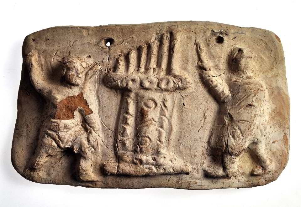 Clay relief showing an ancient hydraulis (water organ) at the center, flanked by two gladiators, each raising an arm as if in a gesture or salute. The figures are dressed in typical gladiatorial attire, and the organ features several upright pipes.