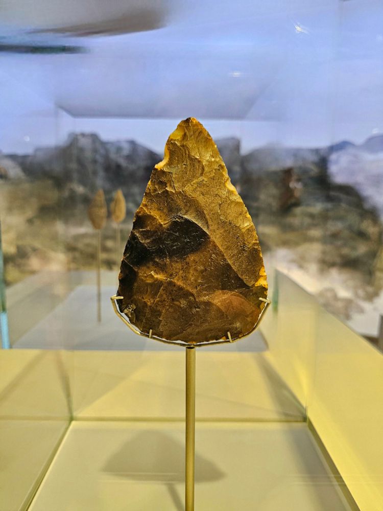 A hand axe made of flint displayed upright in a glass case, with a pointed, triangular shape and a textured, brownish surface. The background shows a blurred image of am outdoor landscape.