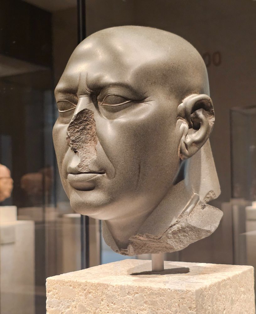 A sculpted head carved from dark stone, portraying a bald male figure with detailed facial, rather realistic features. Despite damage to the nose, ear, and base, the sculpture's lifelike quality remains evident. It is displayed on a light stone pedestal in a museum, with other artifacts visible in the background.