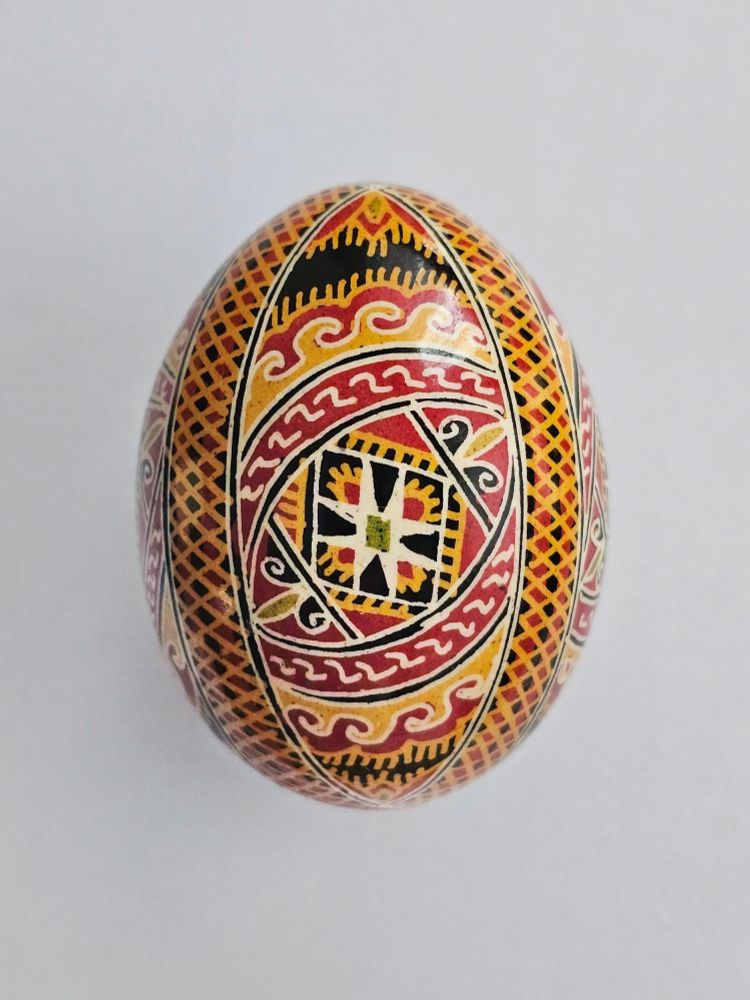  A single intricately decorated egg featuring a symmetrical, traditional design with geometric patterns and vibrant colors including red, yellow, black, and white. The central motif resembles a star or cross, surrounded by wavy and crisscrossed lines in an ornate style,