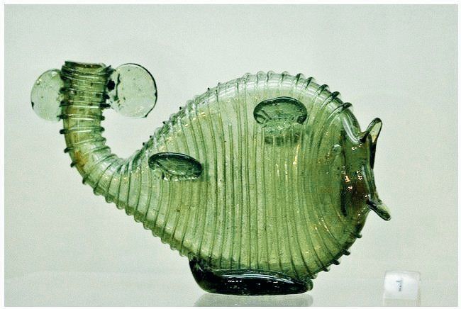 A translucent green glass vessel in the shape of a fish with an oval-shaped body, a curved spout-like tail, and circular fins. The vessel is trailed with thin glass threads.