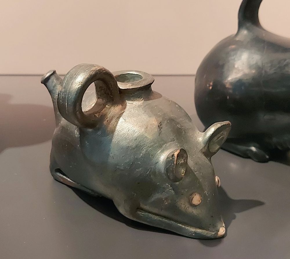 A pottery baby feeding vessel crafted in the shape of a mouse, with a rounded body, pointed snout, and upright ears. The spout resembles a tail, and a circular handle is positioned on the back near the filling opening. 