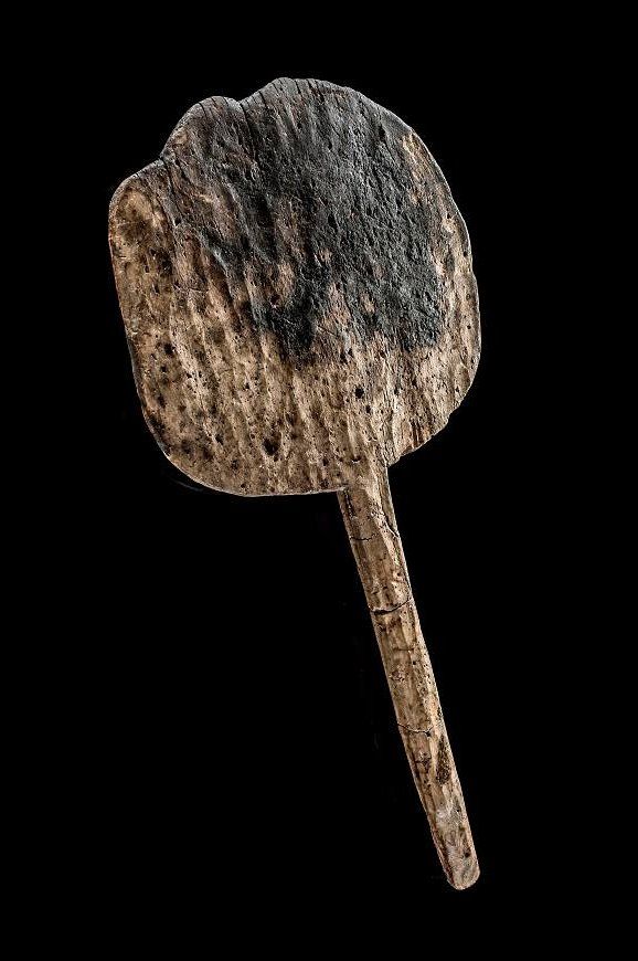 The bread peel is light brownish-gray wood, with visible wood grain and texture, and dark-brown or black markings suggesting charring. The bread peel's head is broad, and the handle extends down from the center of the wide head.
The background is completely black, making the wooden tool stand out as the sole subject. The lighting is even and highlights the tool's shape and surface details without casting deep shadows.