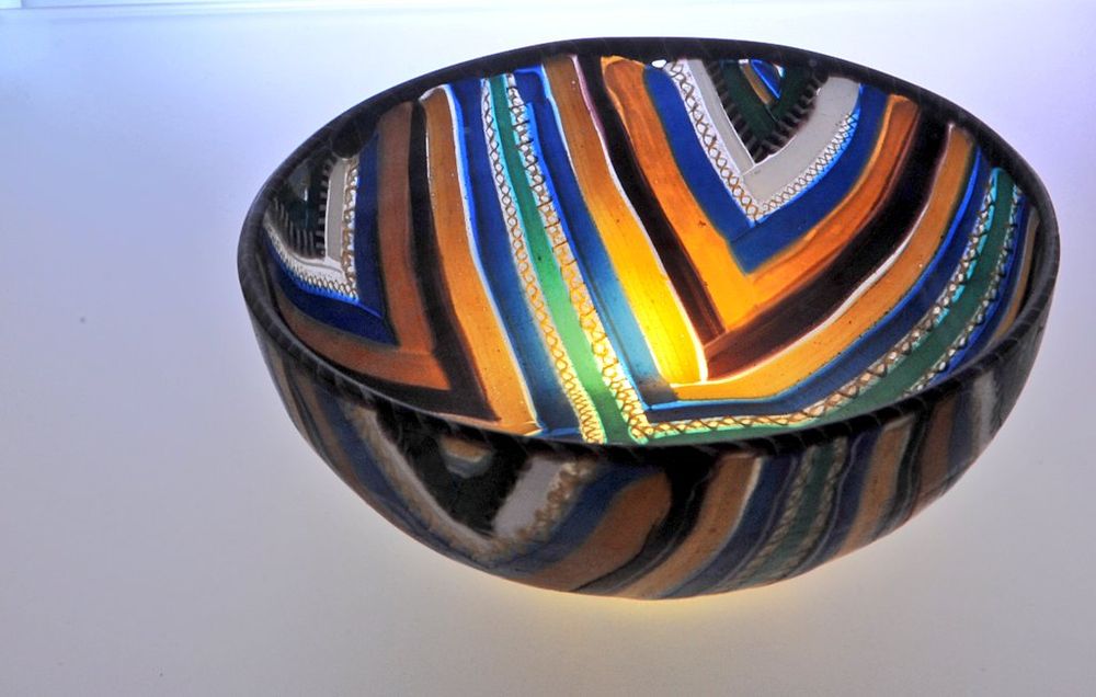 A glass bowl with vibrant, multicolored patterns including blue, orange, yellow, green, and white. The design features bold, angular lines and intricate textures, and the bowl is illuminated , enhancing the colors and creating a glowing effect against a light background.