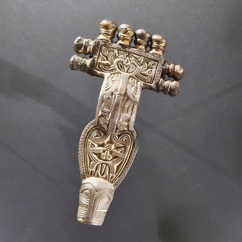 A close-up photo of an intricately designed silver gilt brooch, that features a complex, interwoven design. The top has multiple small, rounded protrusions. The foot ends in a stylized animal head.