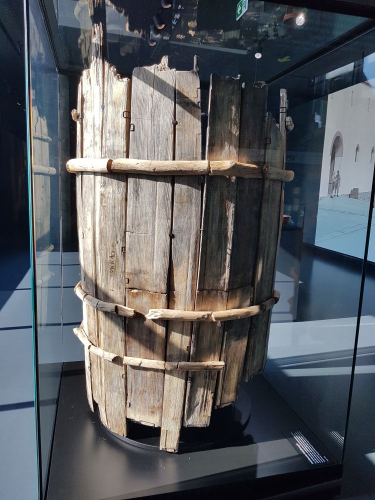 A large, weathered wooden barrel with visible wood grain and several signs of age, such as worn, cracked, and uneven planks, displayed upright in a glass museum case. The barrel appears structurally intact but shows significant wear and damage, with broken or missing sections at the top and bottom edges.