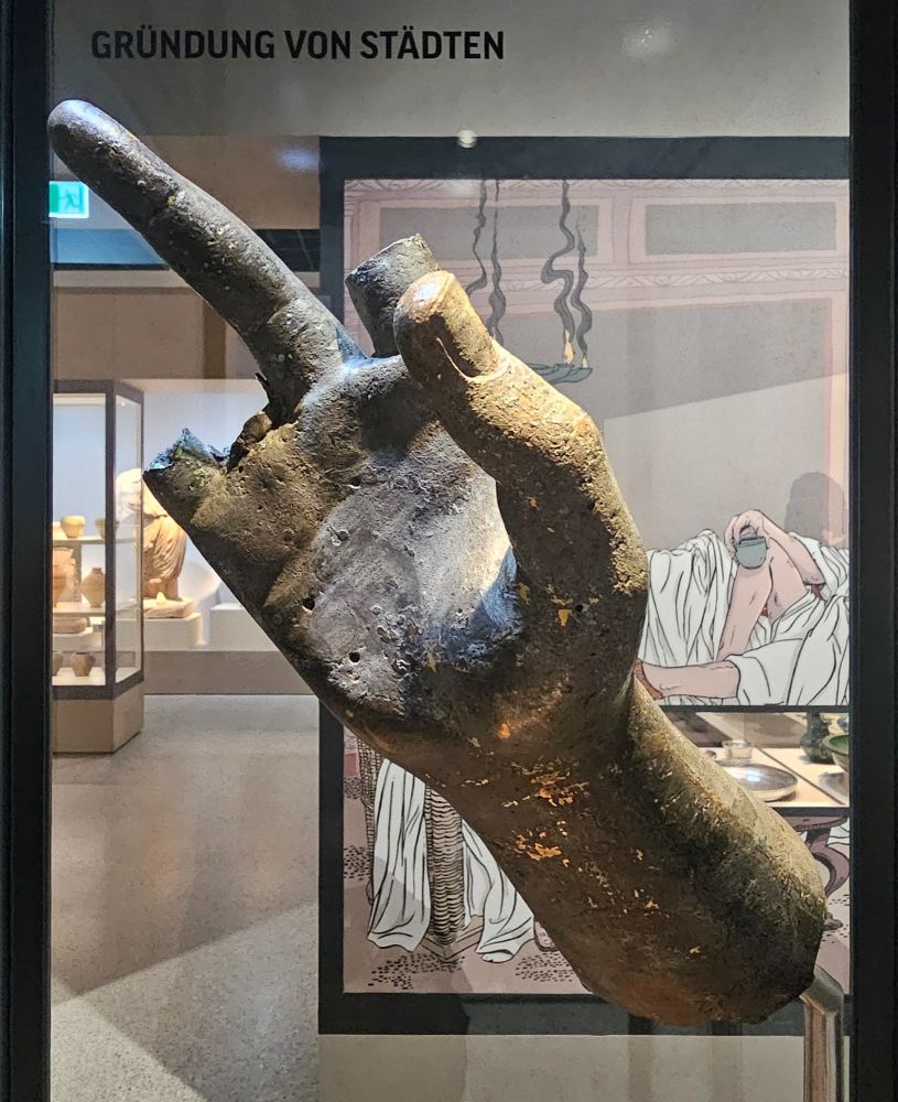 A large bronze hand is displayed in a museum, with its middle finger extended while the other fingers except the thumb are broken off.