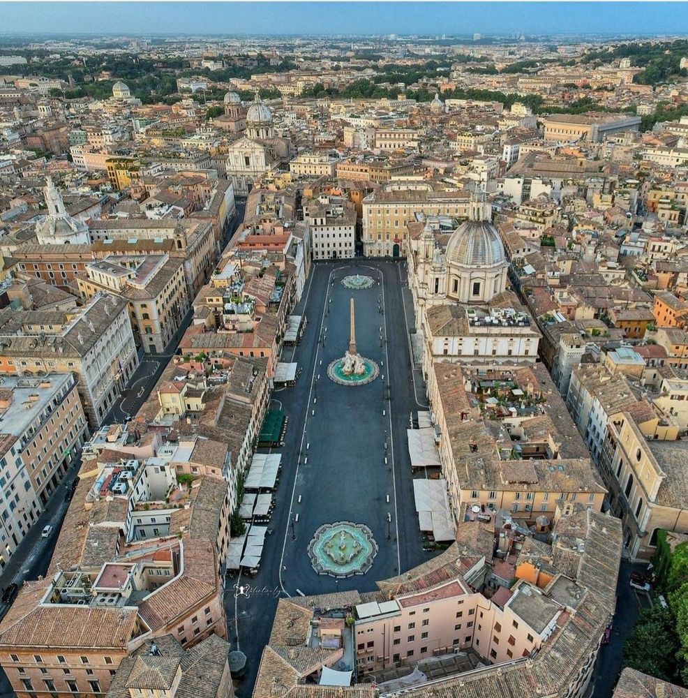 Aerial view of Piazza Navona in Rome, Italy, showing its long, rectangular layout with one rounded end. Built on the site of the ancient Stadium of Domitian, the square features three fountains, including the central Fountain of the Four Rivers with an obelisk, surrounded by historic buildings.