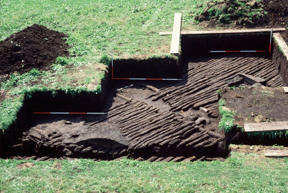 A lawn, squares of which have been dug out until a depth of 30 cm. There are bicolor length indicators (red and white) and, on the flat uneathed bottom: a wooden floor : planks on joists. The wood is black and corroded.