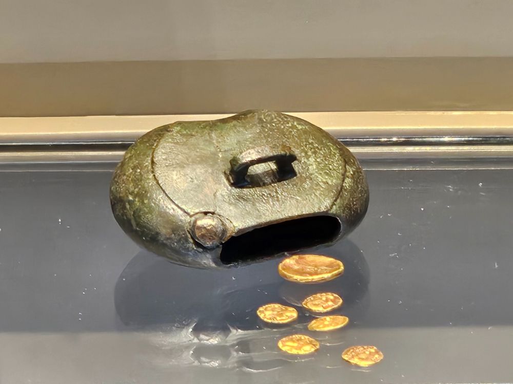 A metal money box made of bronze, rests on display. The oval-shaped container has an open slot where several gold coins have spilled onto the surface below.